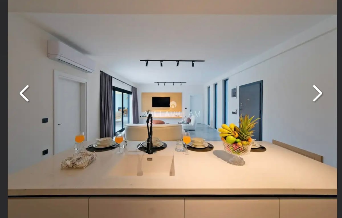 A modern kitchen island is positioned prominently, showcasing a clean countertop with tableware set for four and a fruit bowl. In the background, a spacious living area is visible, featuring large windows, natural light, and a television mounted on the wall.