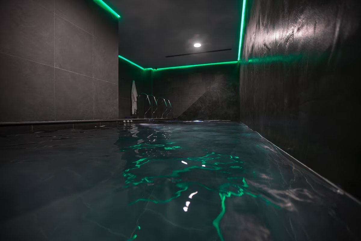 An indoor pool area is presented with sleek dark tiles and modern lighting. Subtle green neon lights create a calming ambiance, reflecting off the water's surface. Comfortable lounge chairs are positioned nearby, contributing to a relaxing environment.