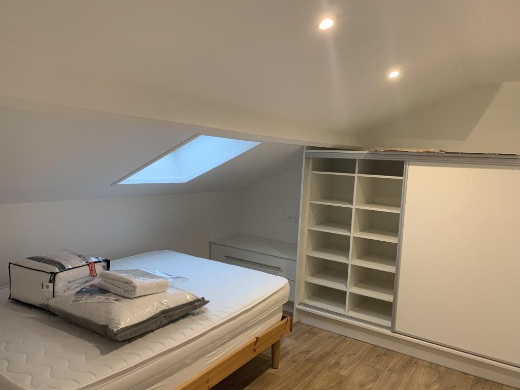 A bright bedroom is presented with a spacious bed partially made, featuring clean linens. A sloped ceiling with a skylight enhances the natural light. Built-in shelving provides storage space, while a minimalist dresser complements the contemporary design.