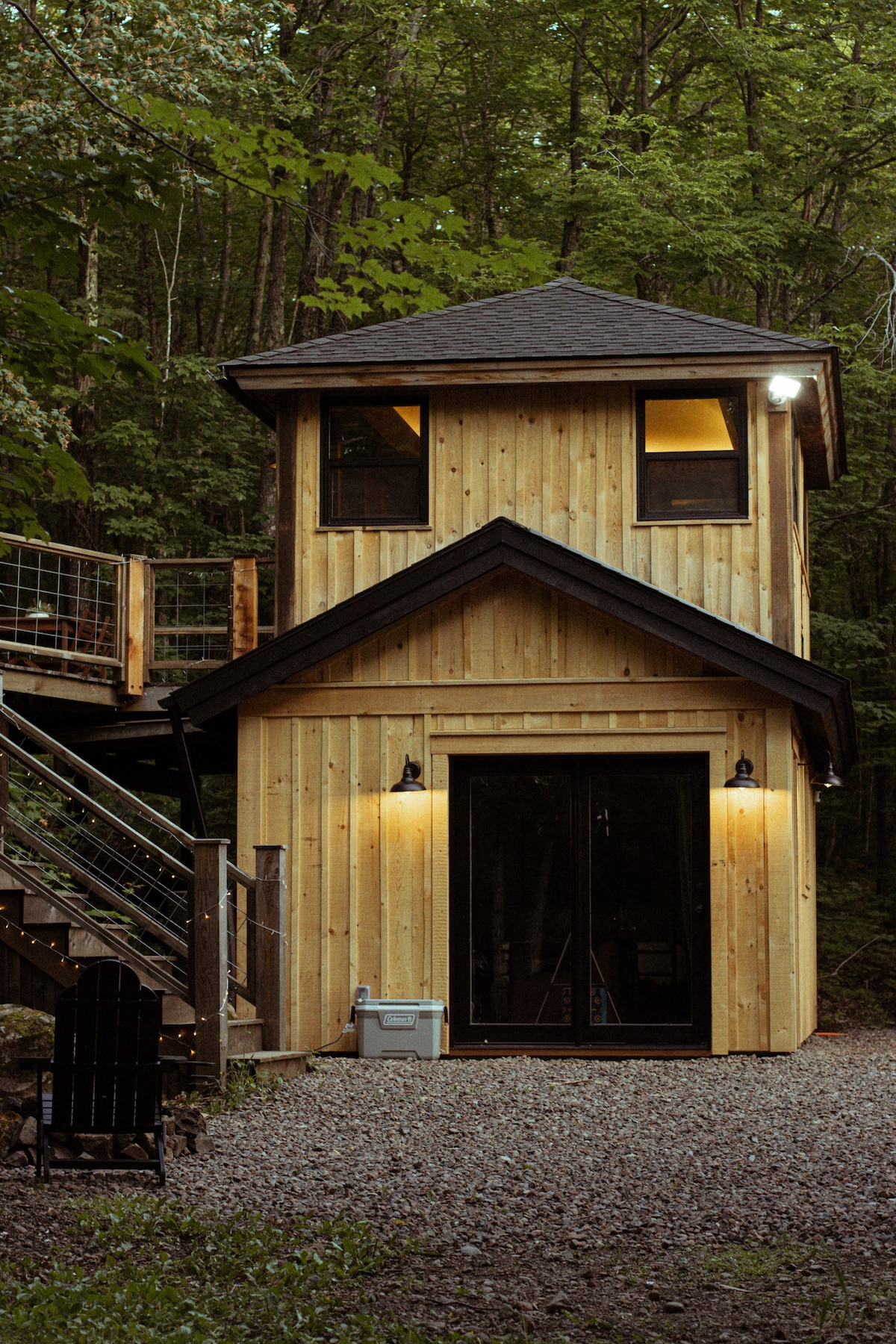 The cabin's exterior is crafted from natural wood, blending seamlessly with the surrounding greenery. A two-story structure features large glass doors at the entrance, providing a view into the interior. Steps lead up to a spacious deck, enhancing outdoor accessibility.