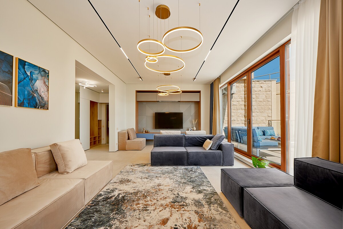 A spacious living area is characterized by large windows allowing natural light, complemented by modern furniture in neutral tones. A unique chandelier comprising circular fixtures offers contemporary lighting. A soft area rug adorns the floor, enhancing the comfortable ambiance without being overpowering.