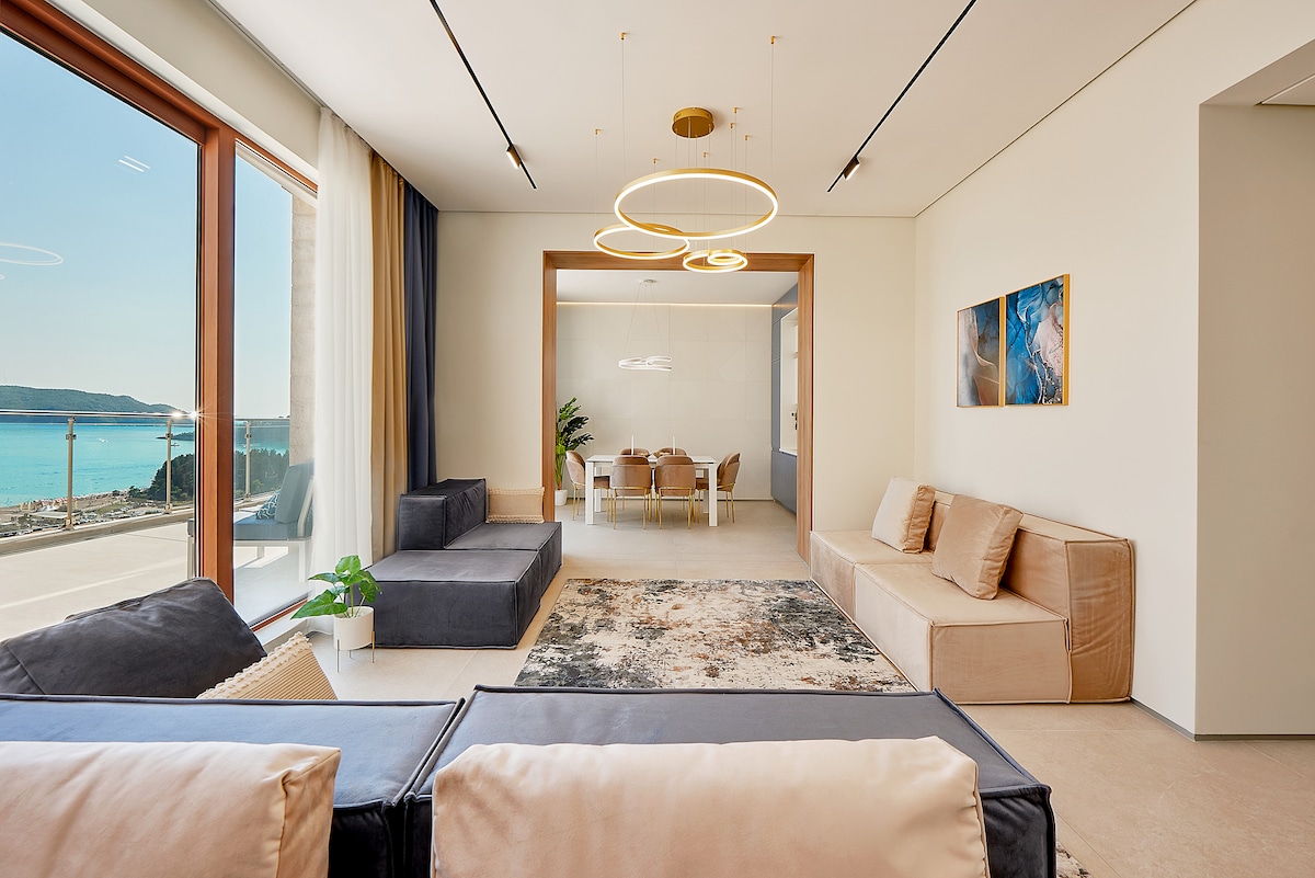 A modern living space features expansive windows offering views of the sea. The arrangement includes a large plush sofa and separate seating areas, with neutral tones and soft rugs enhancing the atmosphere. A dining area is visible in the background, complemented by tasteful artwork.