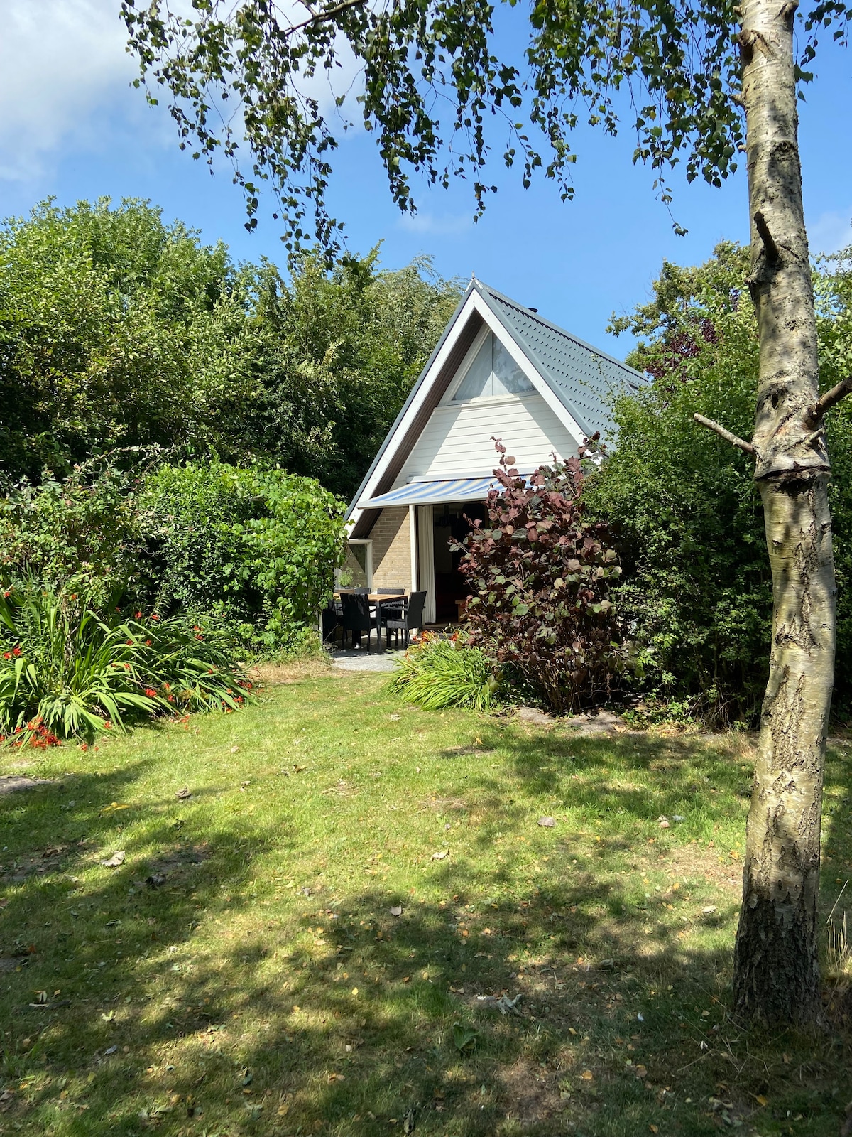 A charming holiday home is set amidst lush greenery, with a sloped roof and a cozy terrace visible. The surrounding garden features a variety of plants, providing a sense of privacy and seclusion. Clear blue skies are seen above, enhancing the inviting atmosphere.