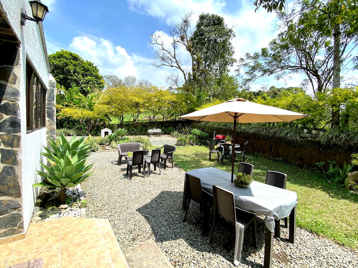 An outdoor space is seen featuring several dining tables and chairs under a large umbrella. Lush greenery surrounds the area, with gravel pathways leading to various seating arrangements. The sky is clear, and sunlight enhances the tranquil atmosphere.
