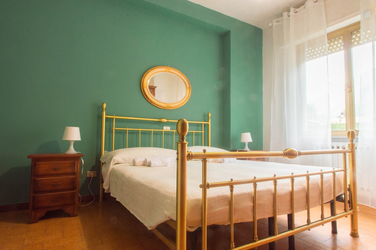 A bright bedroom is presented with a gold-framed bed featuring a white bedspread. A round mirror is mounted above the bed, while two bedside lamps are placed on wooden nightstands. Natural light filters through a window adorned with sheer curtains, illuminating the green wall.