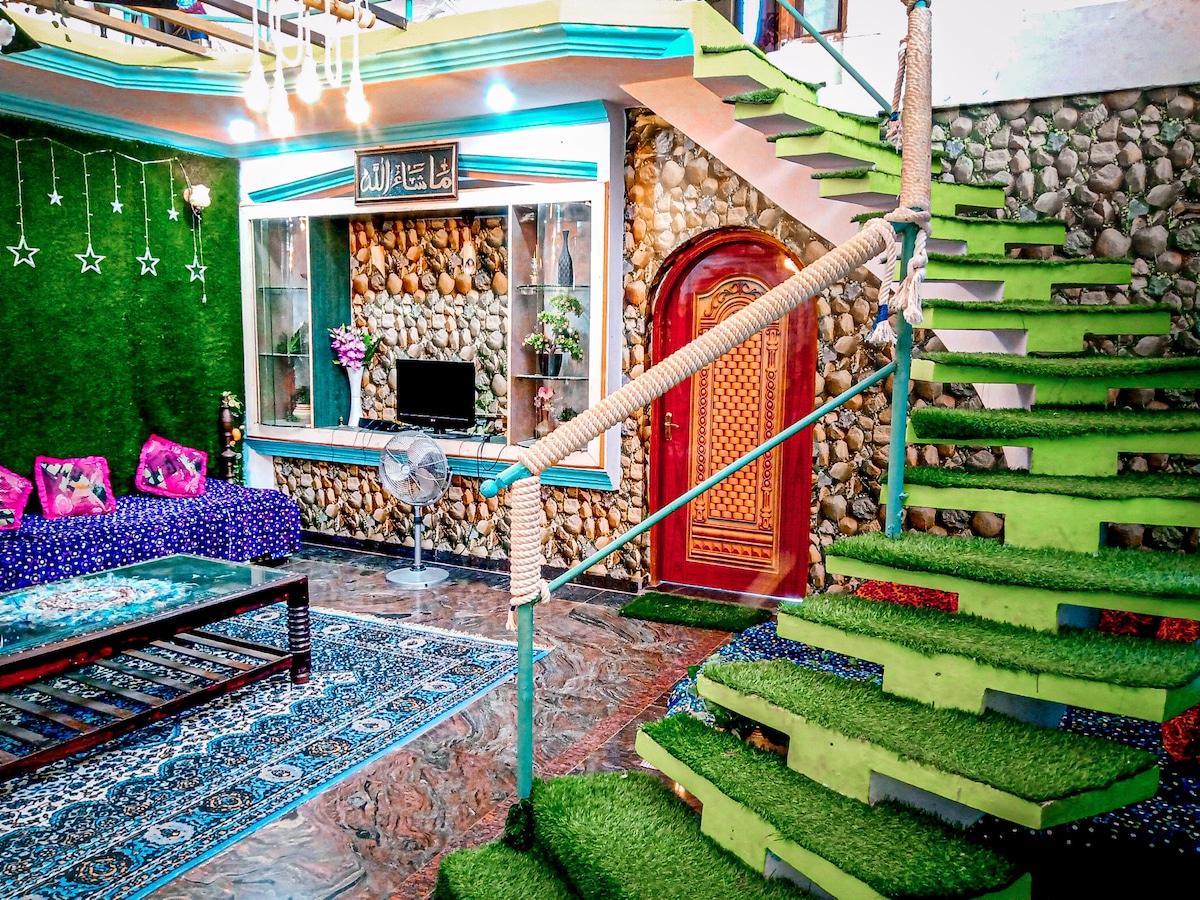 A living area features a combination of stone walls and colorful decorative accents. A plush seating arrangement is set beside a fan and a mounted TV. A staircase with green turf steps leads to the upper level, enhancing the vibrant atmosphere of the space.