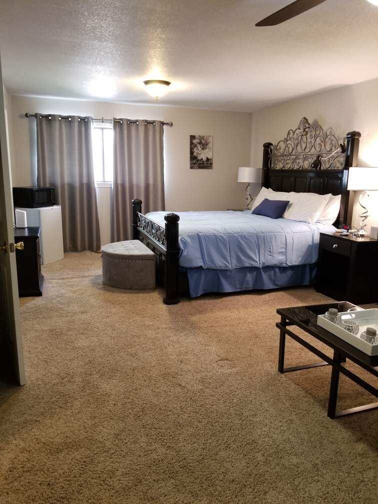 A spacious bedroom features a king-sized bed with a light blue bedspread, complemented by decorative pillows. Two nightstands hold lamps on either side, and a cozy ottoman is positioned at the foot of the bed. A window with sheer curtains allows natural light, while a small refrigerator and microwave are visible in the corner.