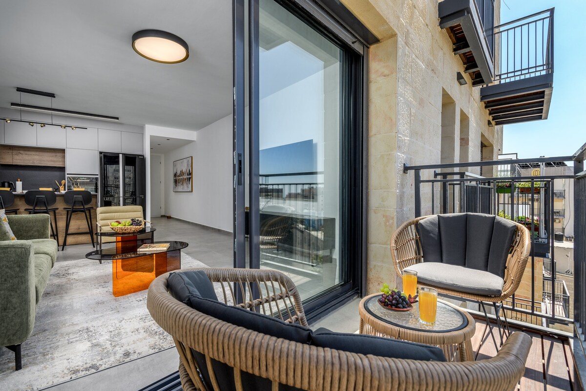 A modern balcony is showcased with comfortable seating, featuring plush chairs and a small table. Sunny drinks and fruit are arranged on the table, with views of the interior, characterized by stylish furnishings and an open kitchen area visible through large glass doors.