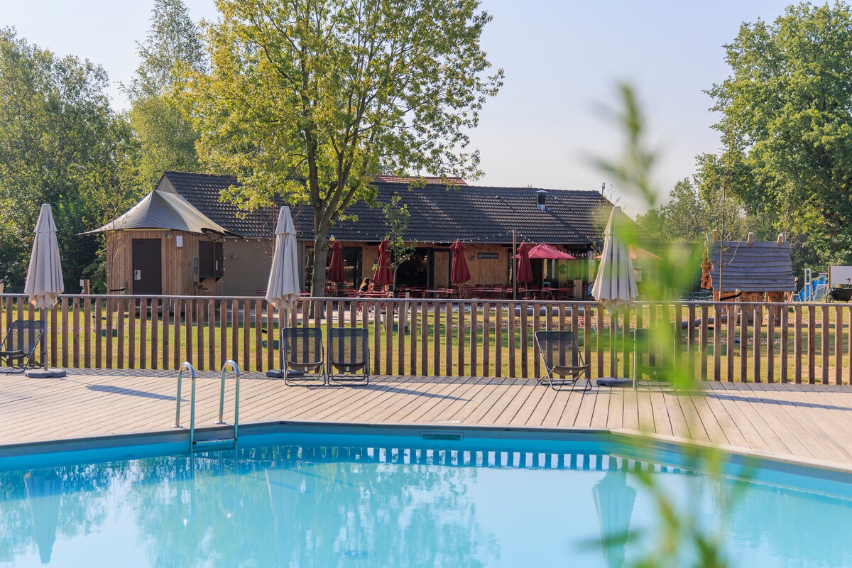 A serene swimming pool is framed by wooden decking, inviting relaxation. Sun loungers are arranged around the pool, with sunshades providing shade. In the background, a welcoming communal area can be seen, featuring seating and dining options beneath a lush canopy of trees.