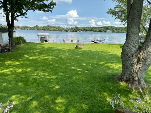 A lush green lawn extends towards a tranquil waterfront, where a boat and seating area are visible. Tall trees provide shade, creating a serene outdoor space. The calm water reflects clouds and distant greenery, enhancing the relaxing atmosphere.