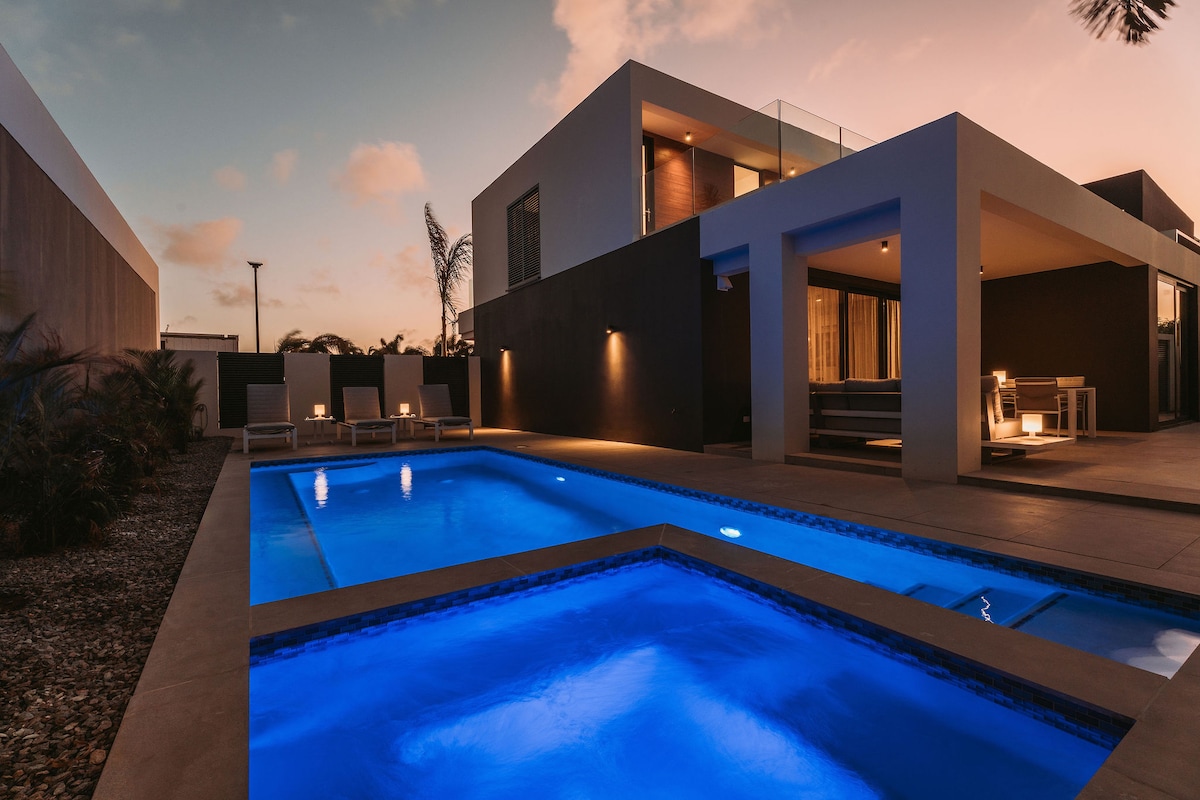 The outdoor area features two sparkling blue pools, surrounded by a spacious patio. Modern lounge chairs are positioned nearby, while the villa showcases a contemporary design, with large windows reflecting the soft evening light.