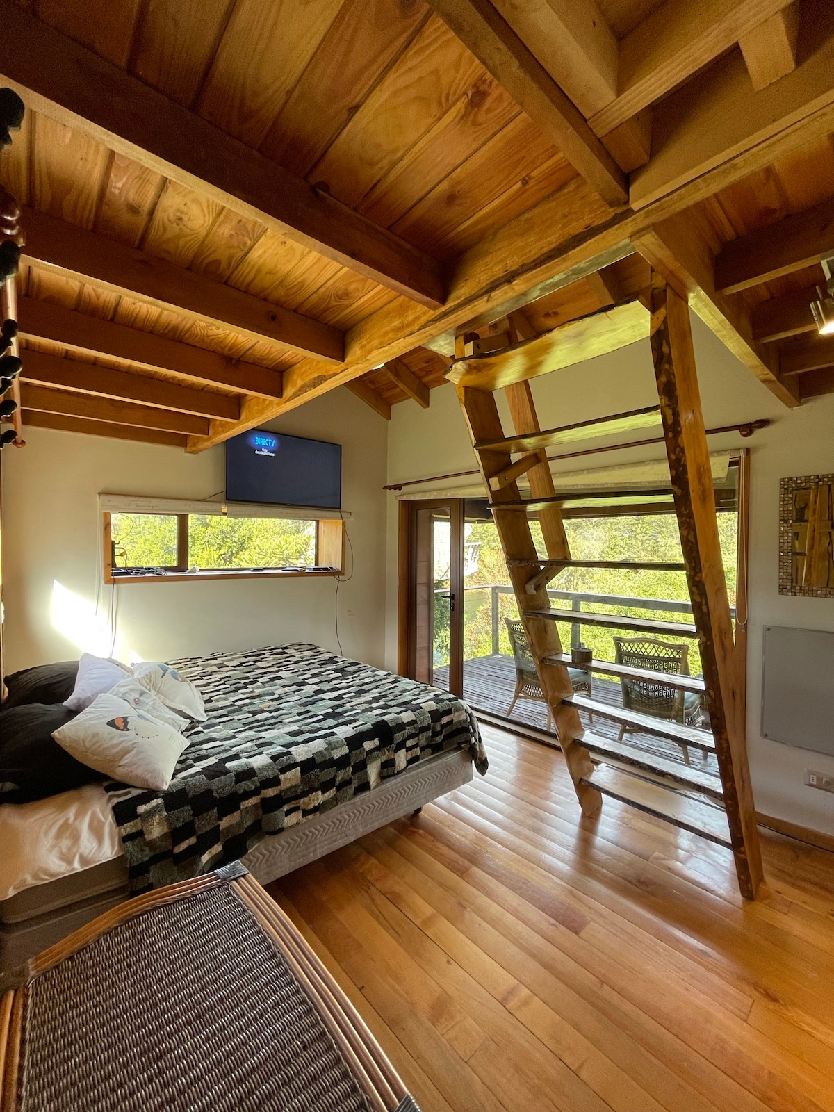 A spacious room features a large bed with a patterned blanket and is complemented by wooden beams overhead. A TV is mounted on the wall, and large glass doors open to a wooden deck, allowing for natural light and outdoor views.
