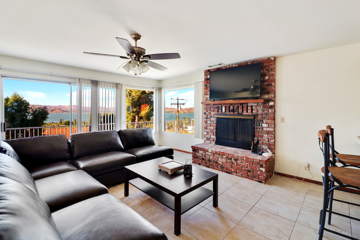 A spacious living area is highlighted by a large sectional sofa and a flat-screen TV mounted above a brick fireplace. Expansive windows frame serene lake views, allowing natural light to fill the room. A dining area with barstools is visible in the background.