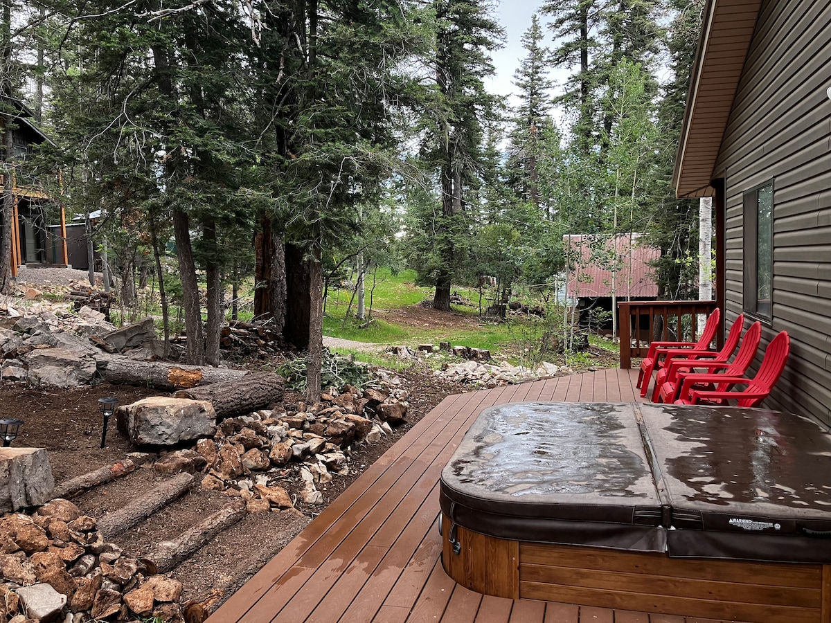 A spacious deck features a hot tub surrounded by wooden decking, with vibrant red chairs positioned nearby. Towering pine and aspen trees create a natural backdrop, while a stone path leads to the wooded area, enhancing the serene outdoor setting.