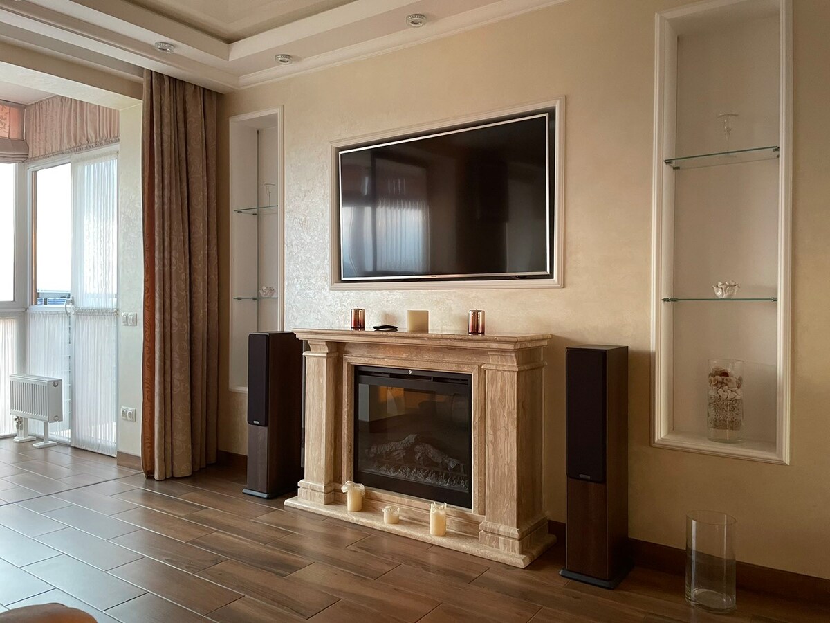 A modern living area is featured, showcasing a marble fireplace beneath a flat-screen television. Shelves on either side hold decorative items. Warm lighting is provided by candles positioned on the fireplace. The space is completed with wood-style flooring and large windows that invite natural light.