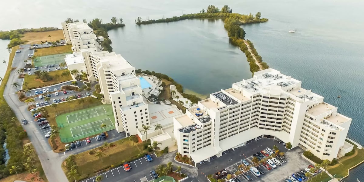Aerial view of a waterfront complex featuring several buildings, a pool area, and tennis courts. Lush greenery surrounds the property, and cars are parked in designated areas. The serene water reflects the sky, enhancing the tranquil vibe of the location.