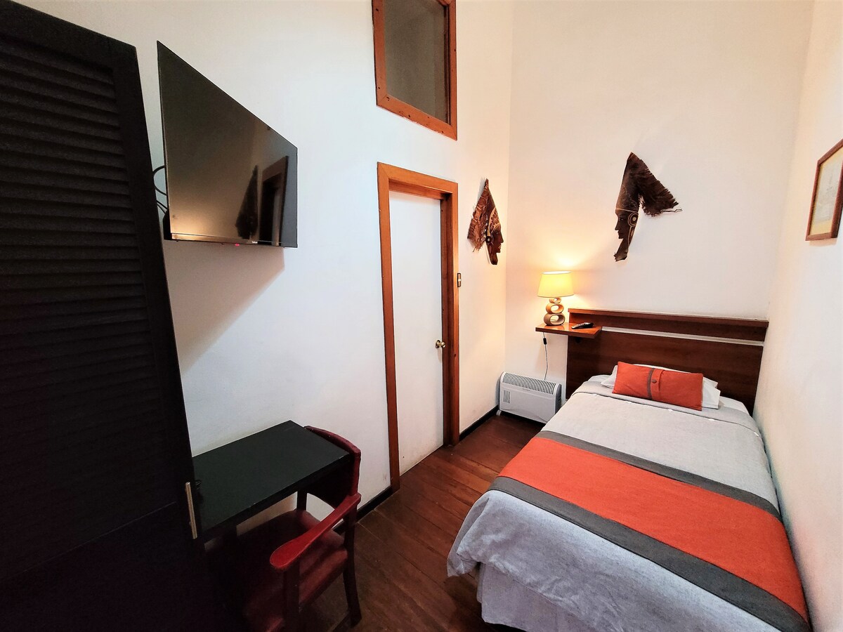 A small room with a bed dressed in gray and orange linens is featured. A wall-mounted TV is positioned above a desk and chair. A door leads to the hallway, and a lamp provides soft lighting on the bedside table. Natural light enters through the high window.