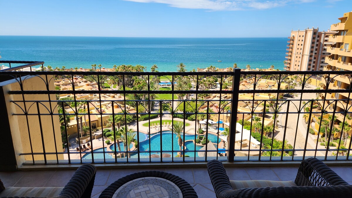 A private balcony overlooks the Sea of Cortez, revealing a stunning view of the resort's pools and lush landscaping. Comfortable seating arrangements offer space to relax while enjoying the tranquil ocean scenery and bright blue sky.