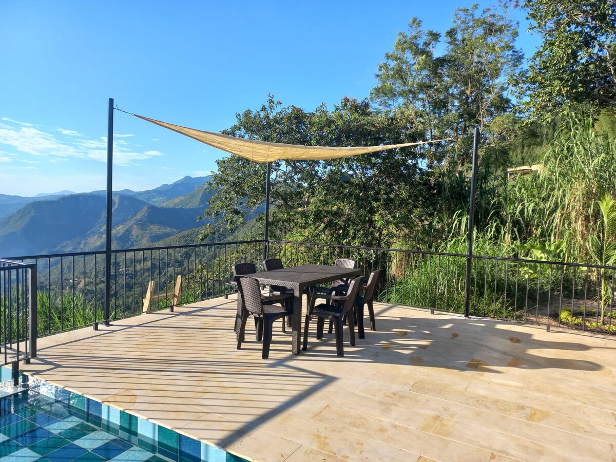An outdoor terrace is equipped with a round table surrounded by six chairs, shaded by a fabric canopy. Lush greenery and mountain views create a tranquil setting, while a swimming pool reflects the clear blue sky.