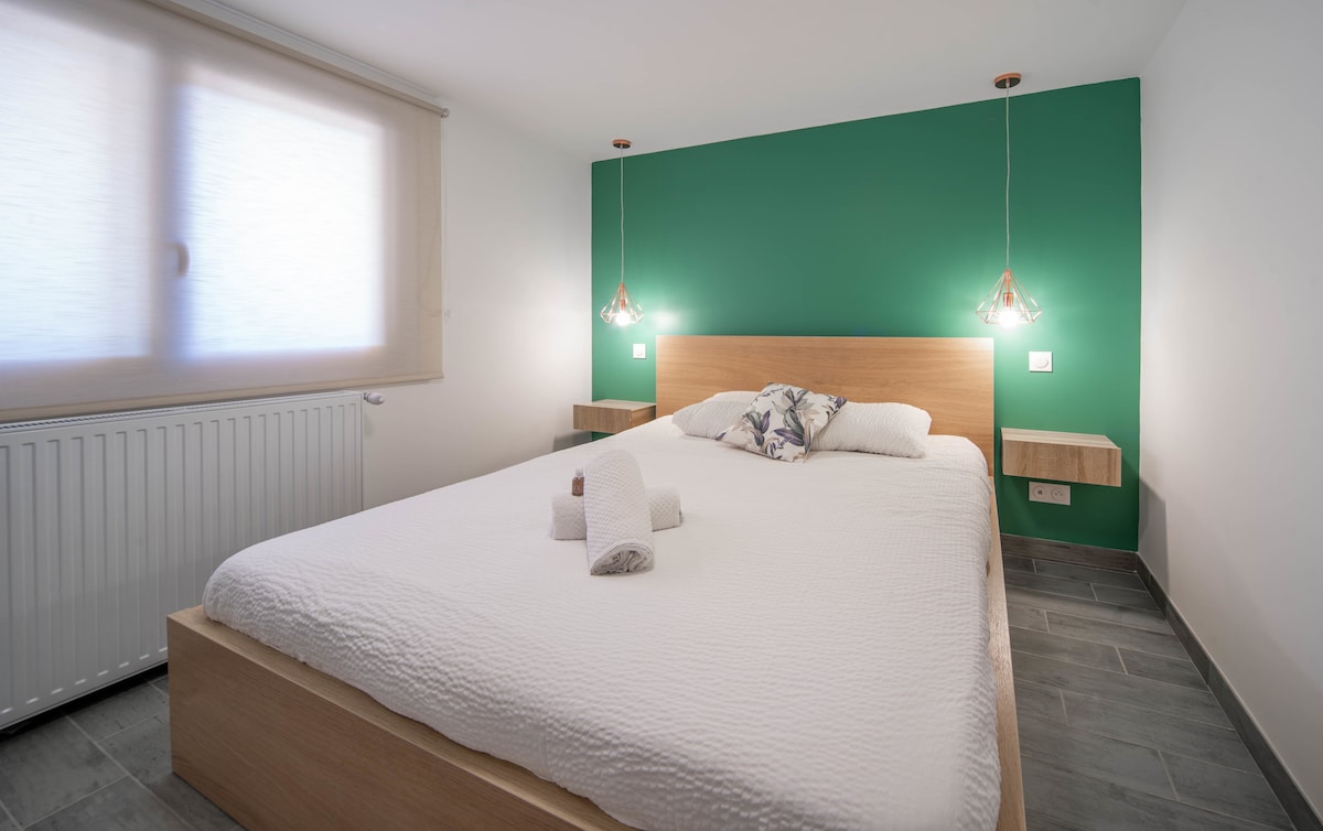 A spacious sleeping area features a comfortable bed with white bedding and decorative pillows. Soft lighting is provided by pendant lights on either side of the headboard. A vibrant green accent wall adds character, while a large window allows natural light to fill the room.