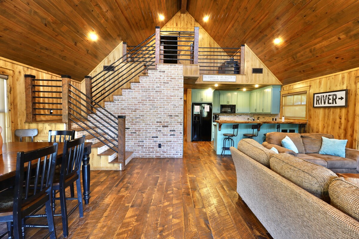 An open and spacious living area features a wooden ceiling and floor, with a staircase leading to the upper loft. A brick accent wall complements the modern turquoise kitchen, which is visible in the background. Dining furniture and a comfortable couch create a welcoming environment.