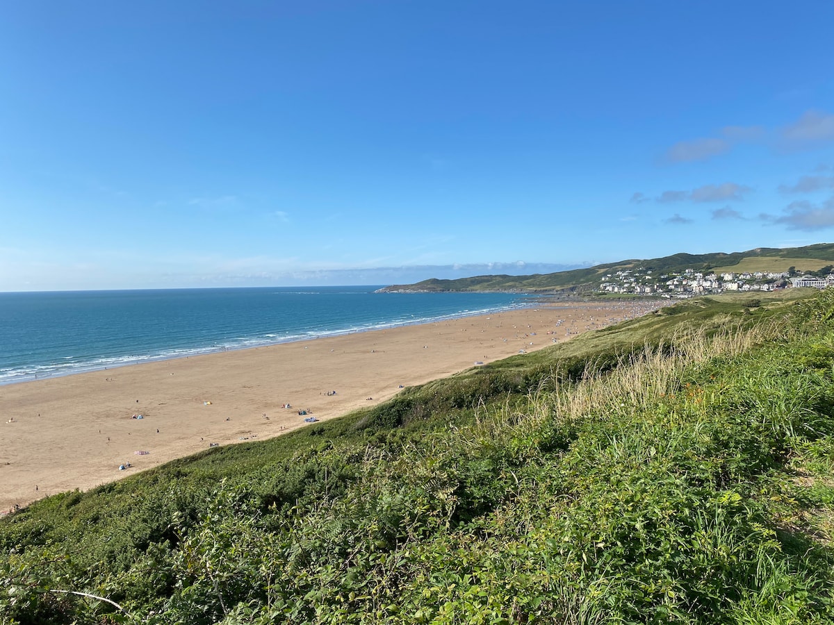 A wide sandy beach stretches along the coastline, meeting the calm blue waters under a clear sky. Gentle waves lap at the shore, while a grassy area leads to the beach. In the distance, coastal houses are visible against the greenery of the hills.