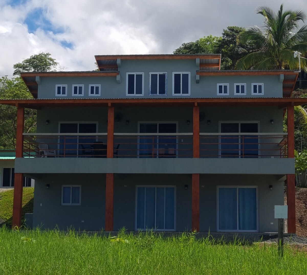 The multilevel home displays a modern design with large windows and glass doors that provide ample natural light. A wide balcony stretches across the front, offering spaces for seating. Lush greenery surrounds the property, enhancing its natural setting.