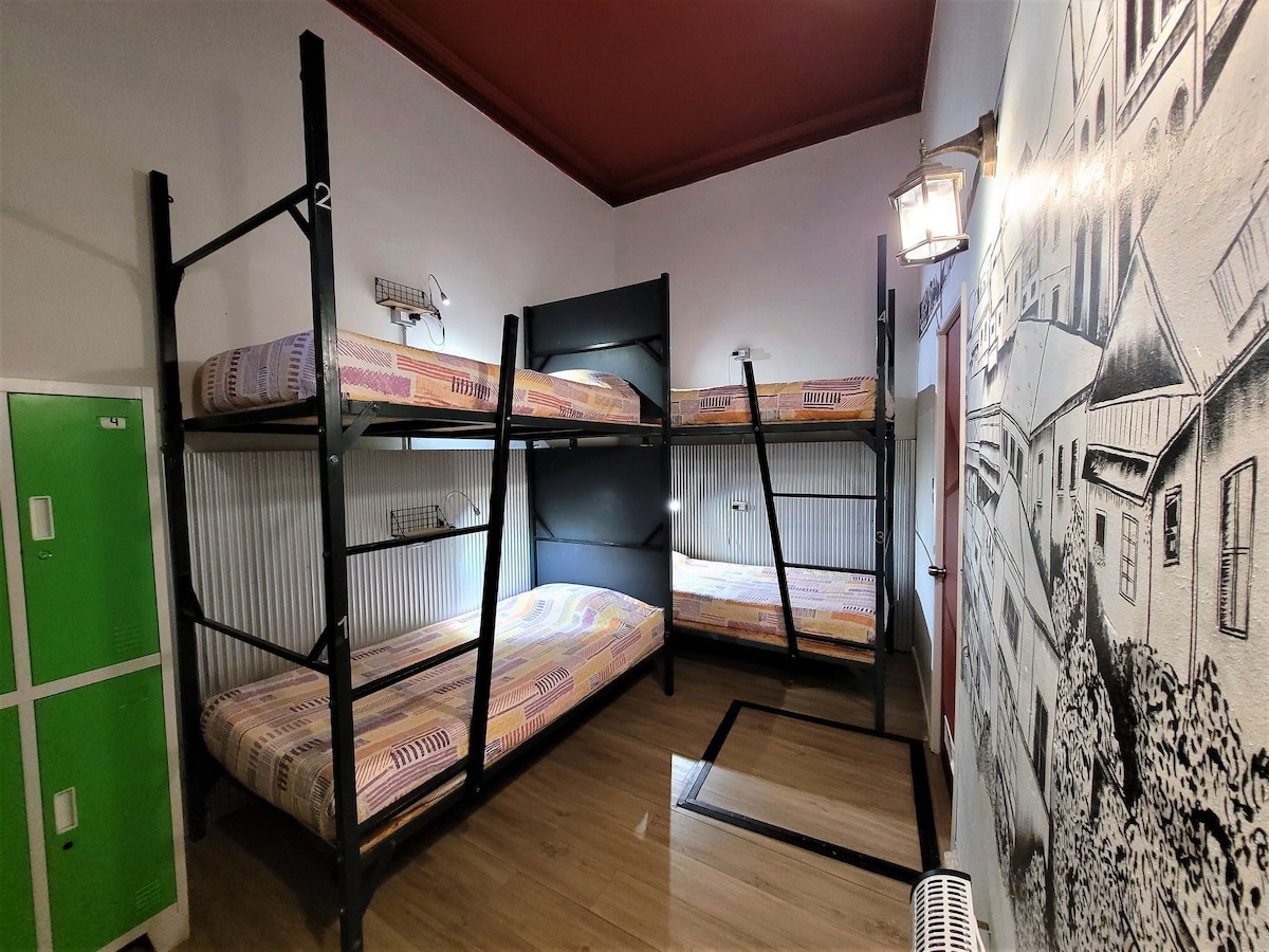 The room features four beds arranged in two bunk beds, providing ample sleeping space. Each bed is equipped with personal lockers, individual reading lights, and power outlets. A wall mural adds a decorative touch to the space, creating a shared accommodation atmosphere.