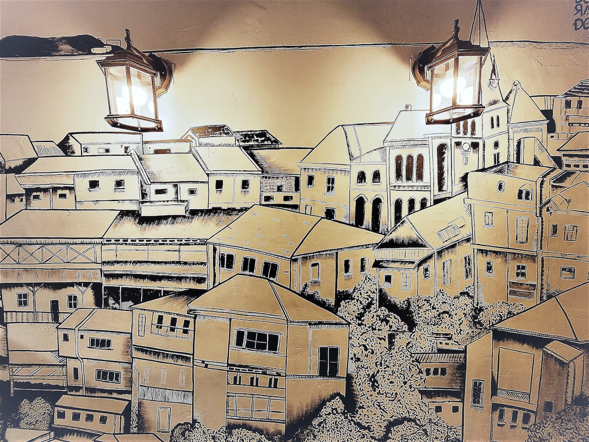 A mural depicting a scenic view of Valparaíso's hillside architecture is illuminated by two vintage-style lanterns. The mural features various buildings with distinct shapes and sizes, showcasing the unique charm of the city with intricate black outlines against a warm, golden background.