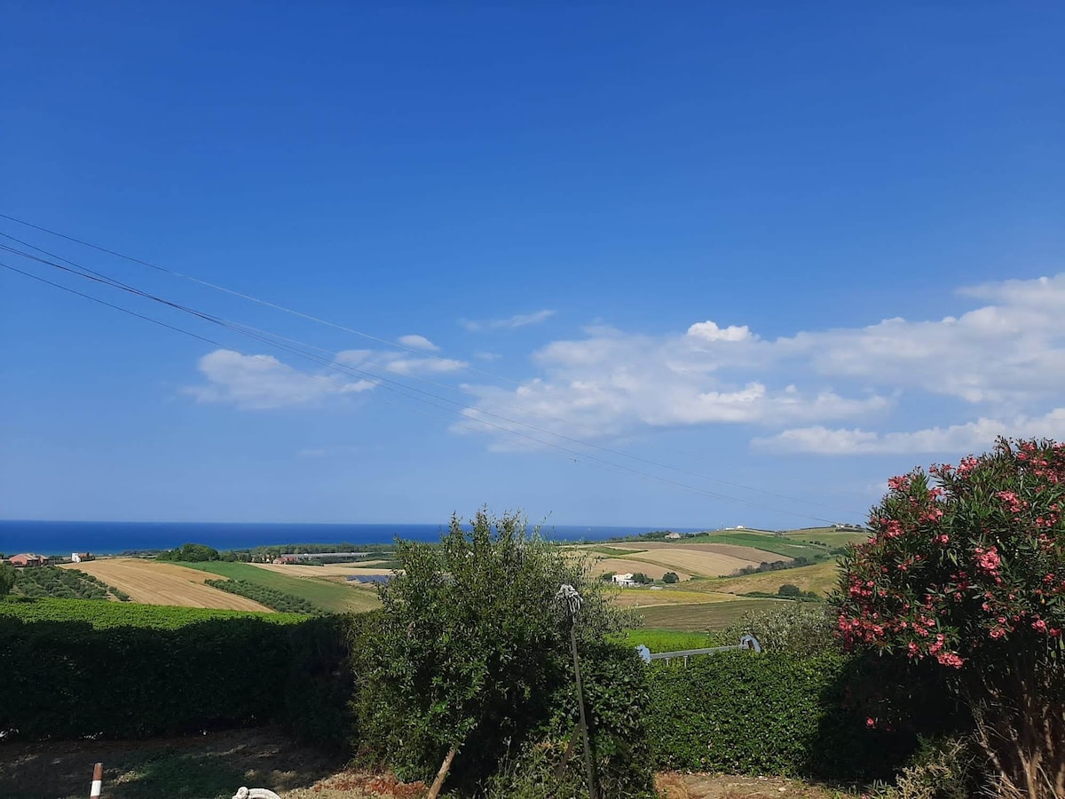 An expansive view showcases rolling hills in vibrant green hues, leading toward a distant sea. A clear blue sky is dotted with soft clouds. In the foreground, lush bushes and a flowering plant add natural color to the serene landscape.