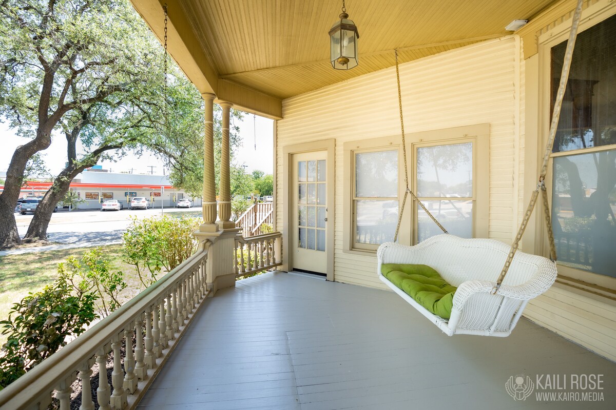 A spacious porch features a white swing adorned with green cushions, providing a comfortable spot to relax. The porch is framed by intricately designed railings, while shaded trees enhance the peaceful outdoor setting. Nearby, the view includes aspects of the surrounding neighborhood.