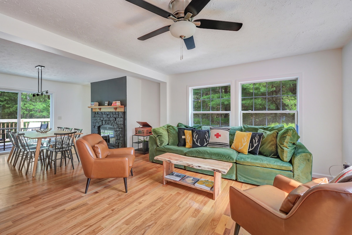 An open-plan living area features an extra deep green couch with colorful pillows, complemented by two brown armchairs. Large windows offer views of the surrounding greenery, while a wooden coffee table sits atop hardwood flooring. A stone fireplace adds warmth to the space.