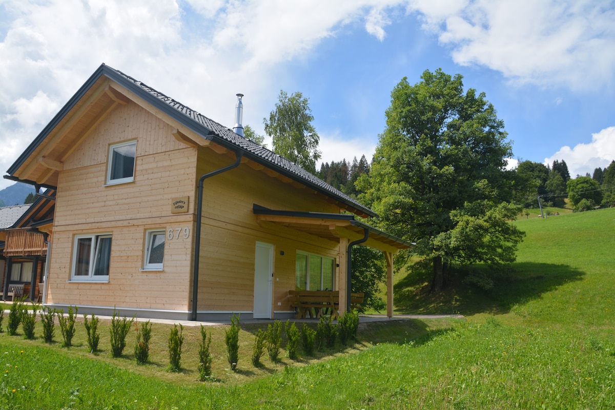 A wooden cottage with a sloped roof is set against a backdrop of green hills and trees. Sunlight illuminates the exterior, highlighting the natural materials. A covered porch area is visible, along with a well-maintained lawn that surrounds the property.