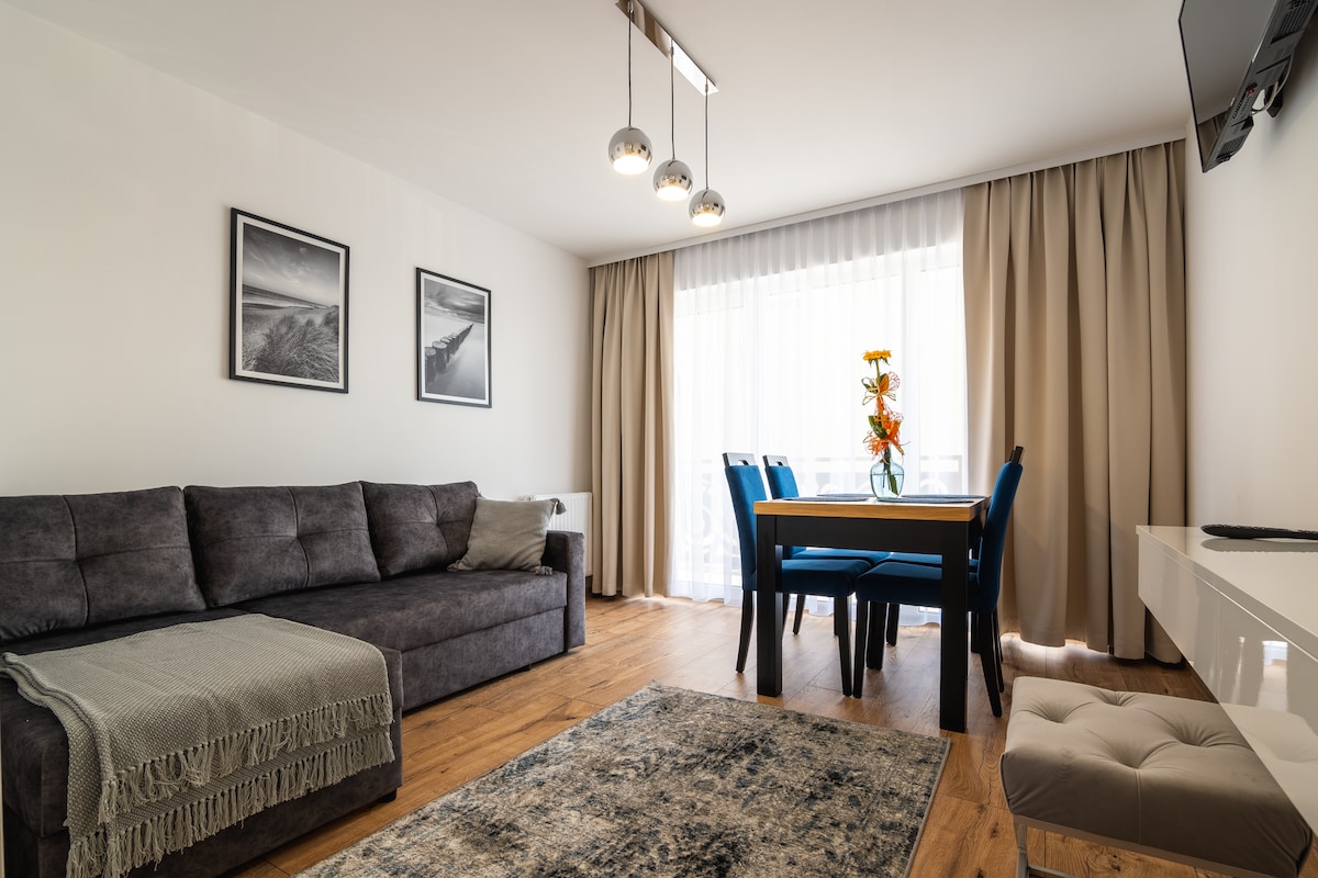 A comfortable living area is depicted with a spacious gray sofa facing a flat-screen television. A dining table with blue chairs occupies the center, and natural light floods through large windows adorned with sheer curtains. Flower arrangements add a touch of color.