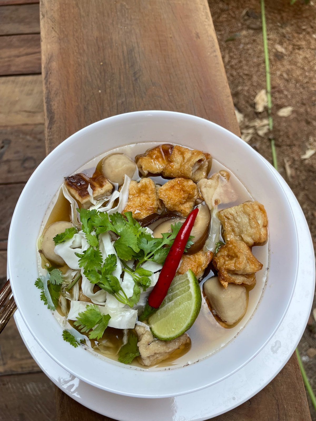 A white bowl filled with a savory broth contains pieces of fried chicken, mushrooms, and noodles. Fresh cilantro and sliced cabbage adorn the top, along with a red chili pepper and a wedge of lime for added flavor.