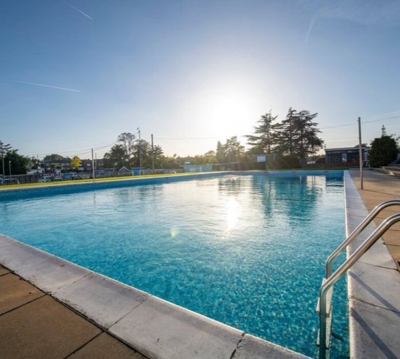 A large outdoor swimming pool is showcased under a bright sun, reflecting shimmering blue water. The surrounding area features tiled decking and metal handrails, with trees and buildings visible in the background, enhancing the open atmosphere.