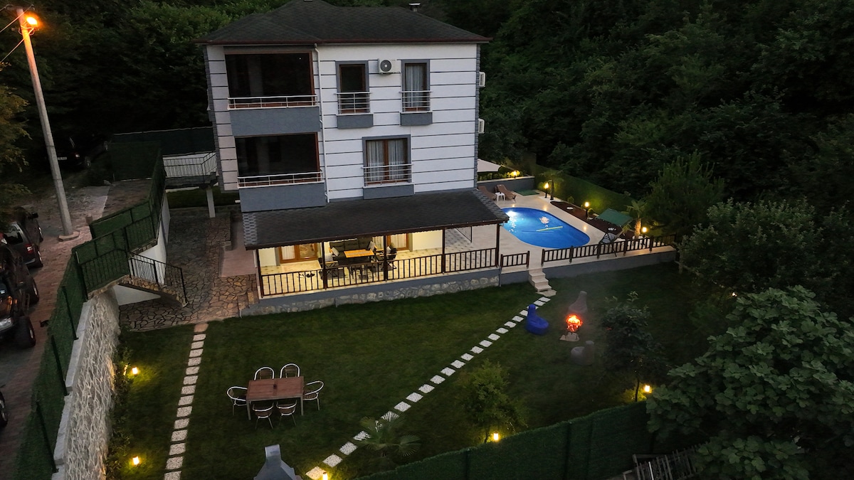 A two-story villa is set amidst lush greenery, featuring a private pool surrounded by a well-maintained lawn. Outdoor seating areas are visible, along with a stone pathway leading from the house to the pool. Subtle lighting highlights the outdoor space during the evening.