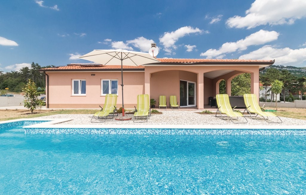The exterior of the house features a light pink facade, complemented by a covered terrace and a private pool. Several lounge chairs are arranged around the pool, accompanied by a large umbrella. The sky is partly cloudy, with greenery in the background.