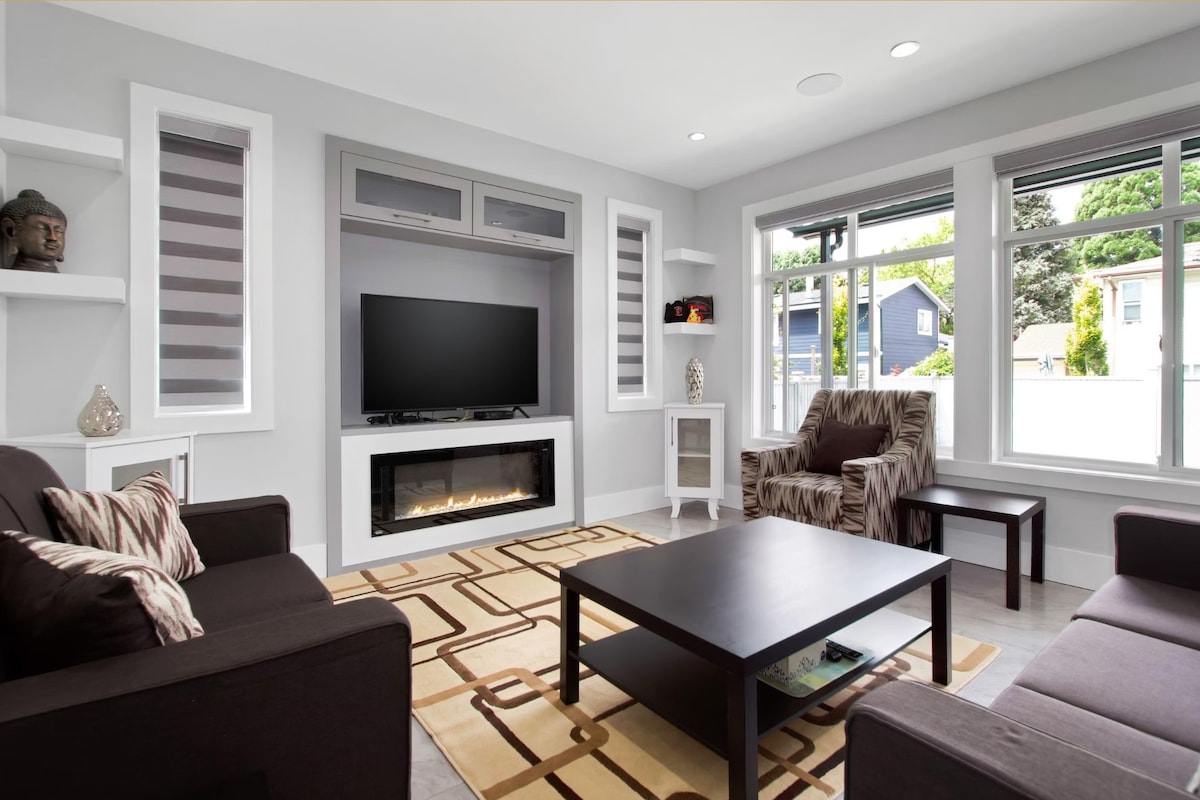 A modern living area features a comfortable brown sofa and an accent chair, with a sleek coffee table at its center. Natural light filters through multiple windows, enhancing the space. An entertainment unit houses a television above a contemporary electric fireplace.
