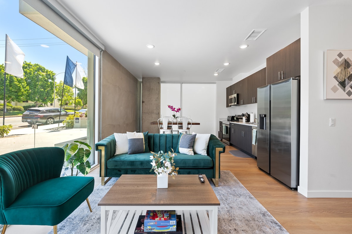 A modern open-plan living space features a plush green sofa and a coffee table with decorative flowers. Floor-to-ceiling windows illuminate the area, creating a bright ambiance. The fully equipped kitchen is visible in the background, showcasing stainless steel appliances and a dining area with a table set for four.