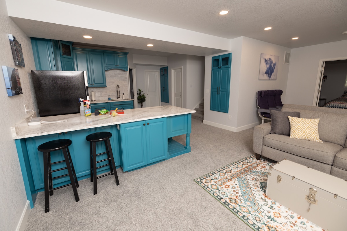 A modern kitchen area features bright blue cabinetry and a spacious countertop. Two black stools are placed at the counter, with snacks arranged on the surface. A cozy living area with a gray sofa is positioned nearby, complemented by a decorative rug and a vintage trunk.