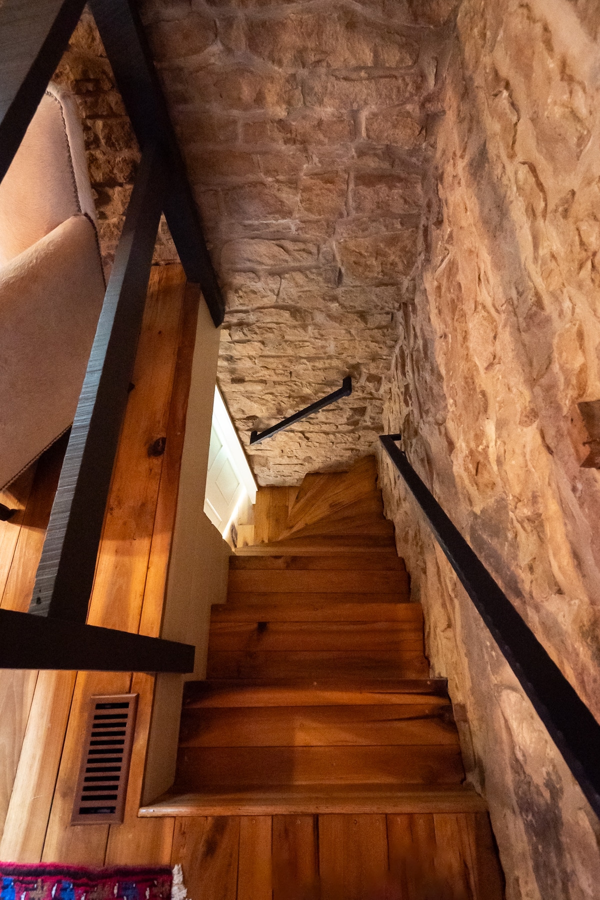 A narrow staircase with wooden steps leads downward, flanked by a stone wall on one side. The natural texture of the stone contrasts with the smooth wood, while a black railing provides support. The descent opens to a light-colored doorway at the bottom.