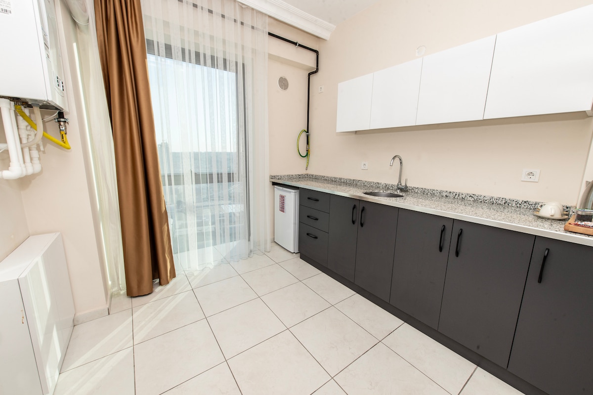 A modern kitchen area features sleek cabinets in a two-tone design. A granite countertop extends along the wall, complemented by light-colored tiles. Natural light streams in through a large window covered by sheer curtains, enhancing the spacious feel of the room.