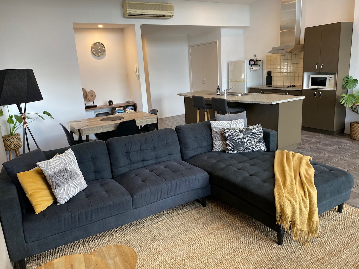A modern living area features a dark sectional sofa adorned with colorful cushions and a soft throw blanket. A round wooden table is visible, along with a kitchen area that includes a sleek countertop and appliances. Soft lighting enhances the open-plan design.