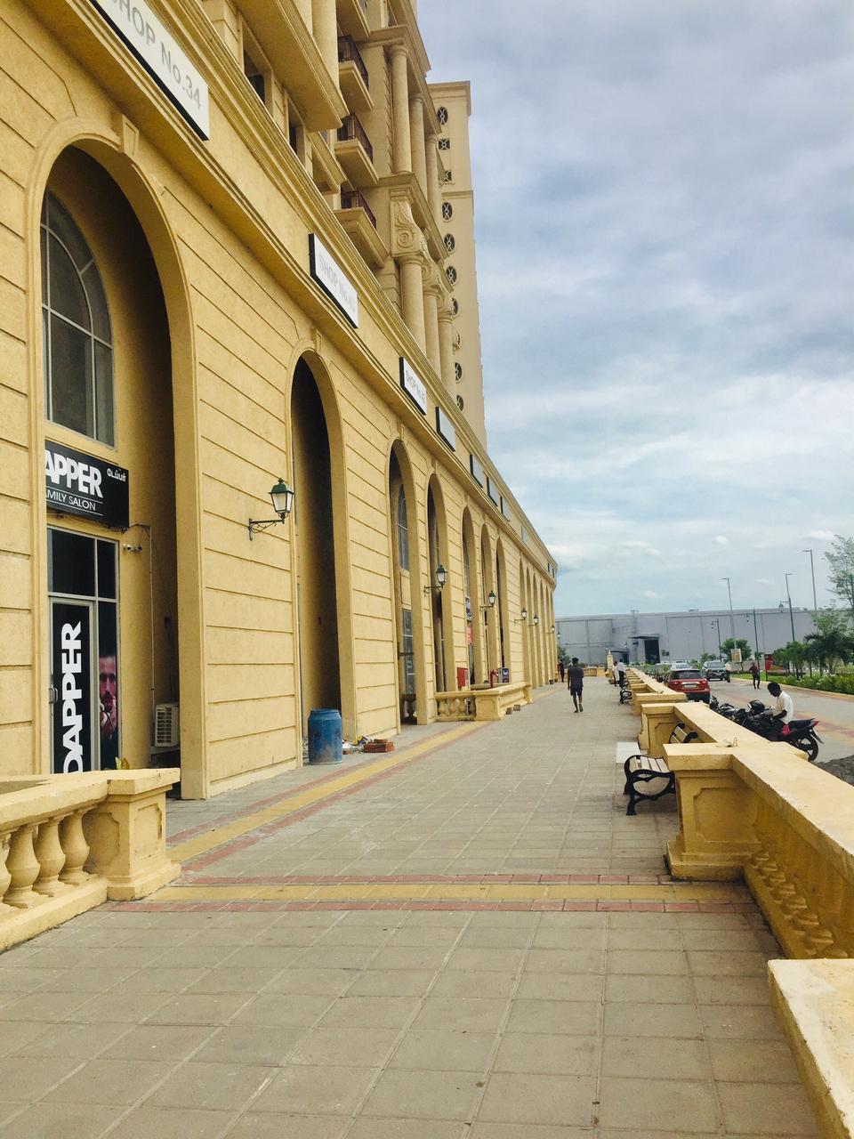 A wide pathway is seen alongside a building featuring arched windows and an elegant façade. Benches are placed along the walkway, inviting visitors to rest. A clear sky and trees are visible in the distance, creating an open and spacious environment.