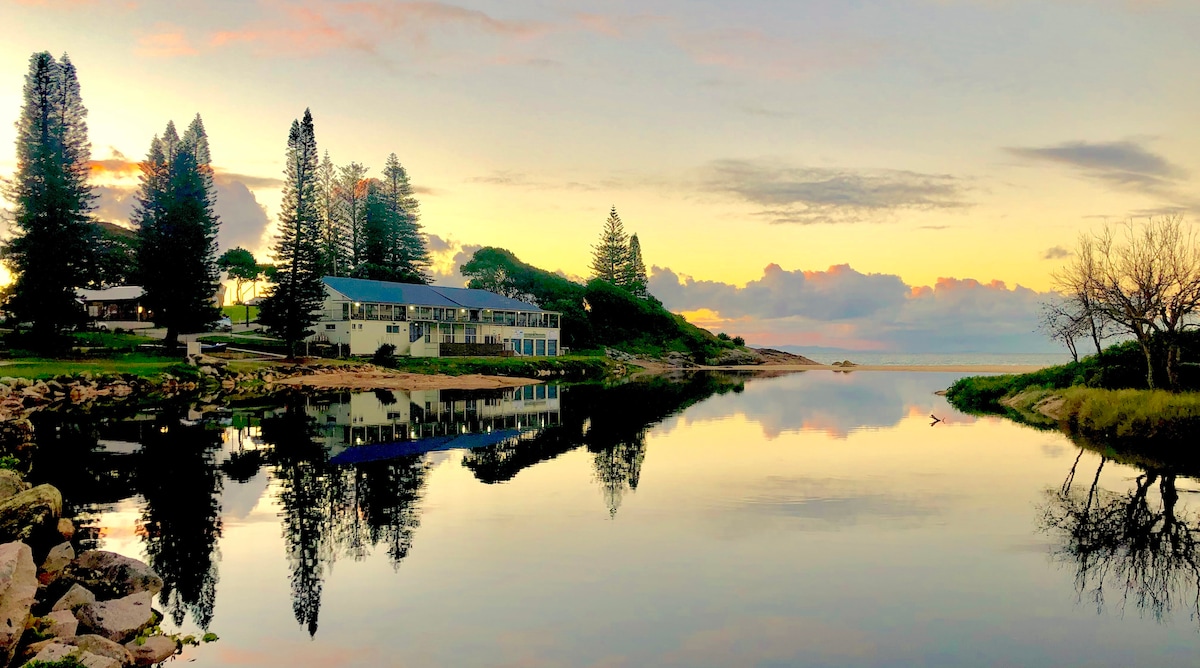 A calm body of water reflects the colorful hues of the sky at dusk. Lush trees line the shore, complementing a single-story building nestled along the waterfront. The tranquility of the scene is enhanced by gentle ripples in the water.