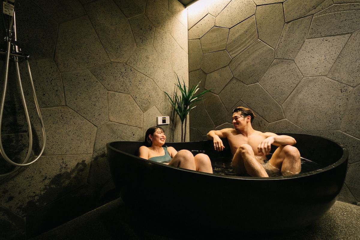 An intimate bathing space is depicted, featuring a smooth, dark stone bathtub surrounded by textured gray walls. A minimalist plant adds a touch of greenery, while a shower fixture is installed nearby, enhancing the serene atmosphere.