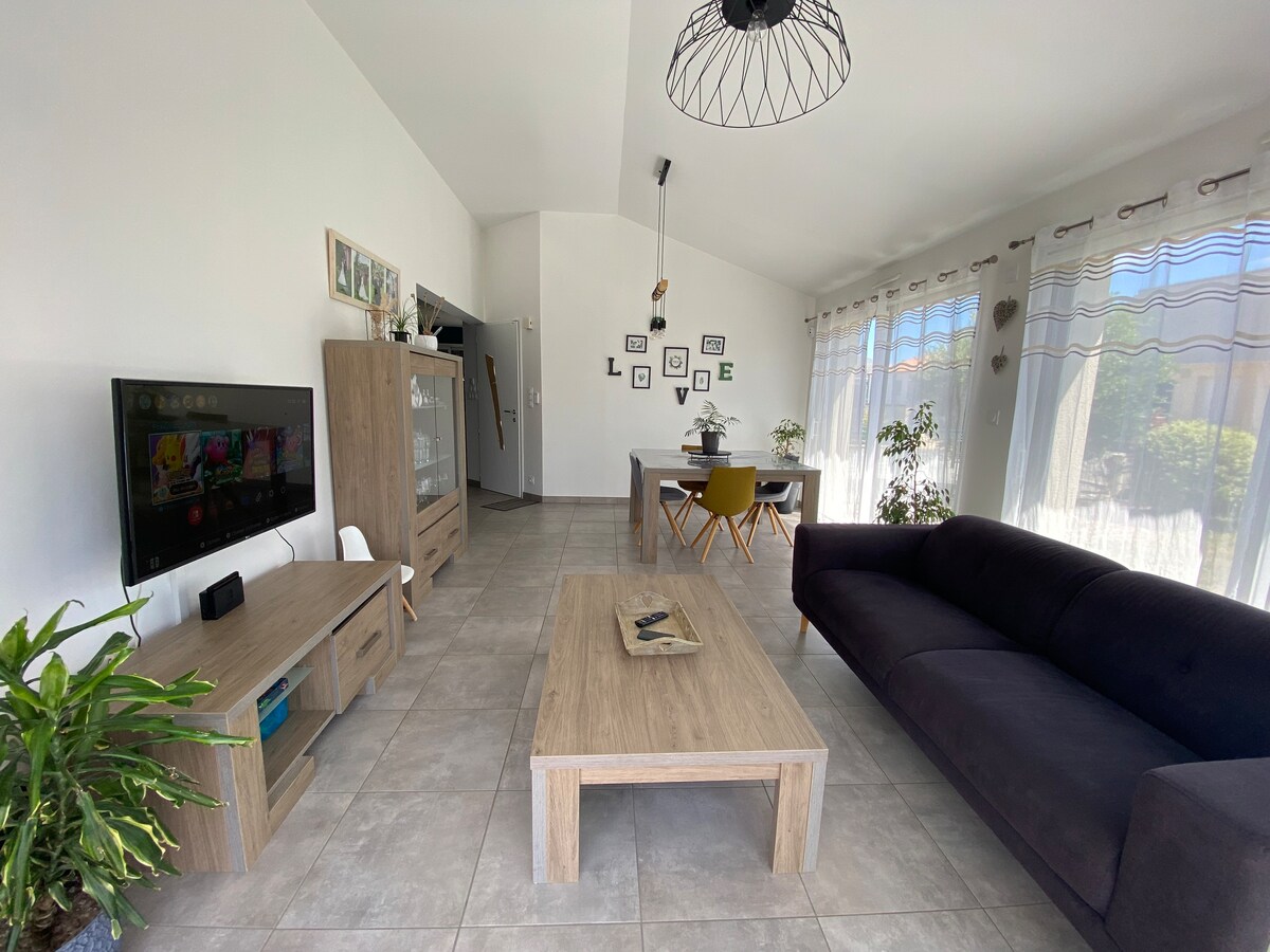 A spacious living area features a dark sofa and a wooden coffee table. A television is mounted on the wall, and a dining table with colorful chairs is positioned nearby. Large windows allow natural light to enter, creating a bright and airy atmosphere.