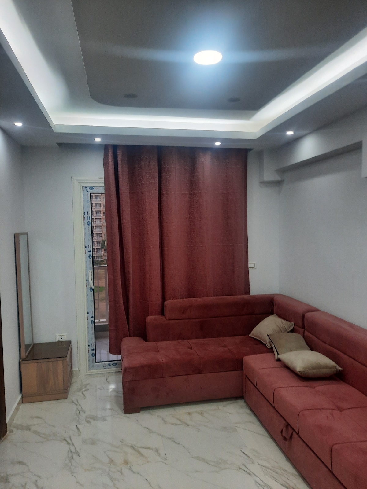 A cozy living area features a deep red sectional sofa with soft cushions, positioned against a light grey wall. Natural light enters through a glass door adorned with sheer curtains, while a rectangular mirror stands beside a small wooden side table.