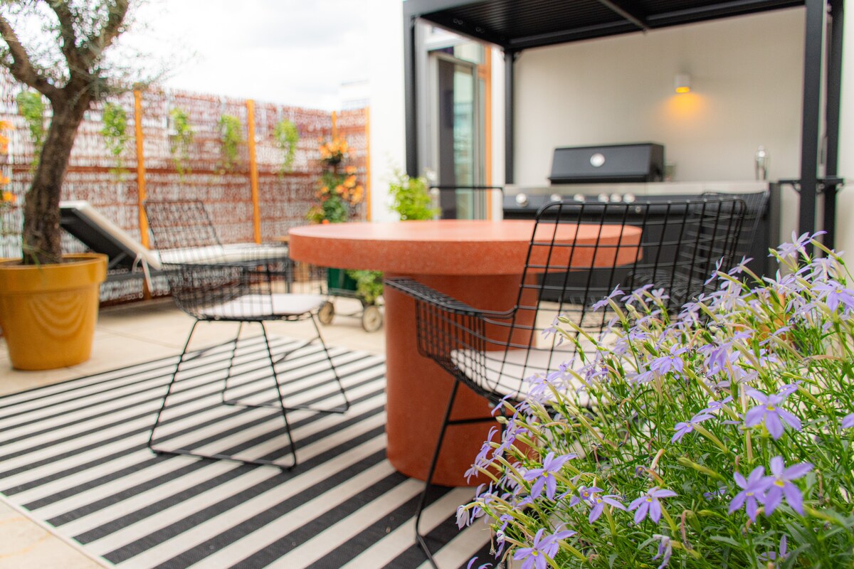 A charming outdoor space features a round, red table surrounded by two wire chairs. A gas grill is located under a shaded area, with a striped rug beneath. Lush purple flowers bloom in foreground, enhancing the inviting atmosphere of the terrace.